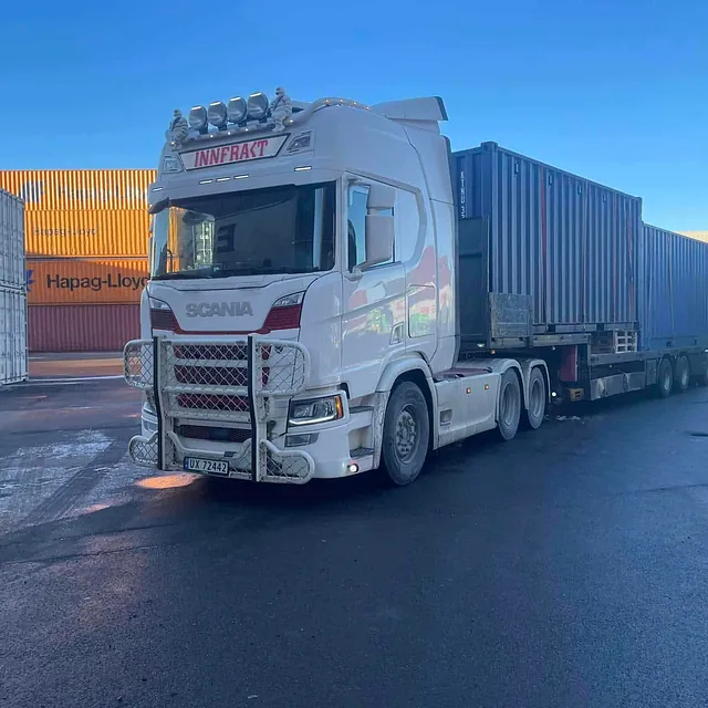 Hvit lastebil frakter blå containere i containerhavn.