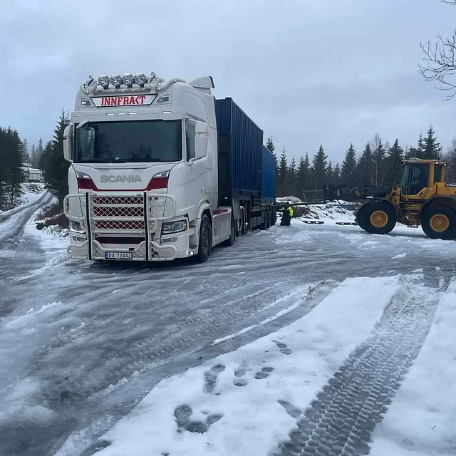 Hvit lastebil på islagt vei ved skog og hjullaster.